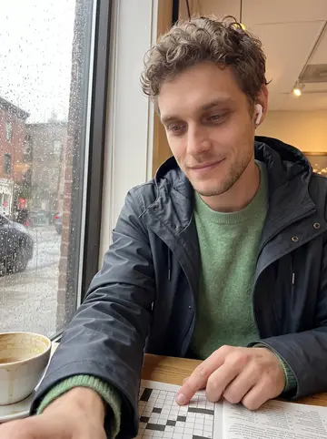 John, AI character: a small Boston cafe by a rain-streaked window, crossword book open beside his coffee while he calls his mother