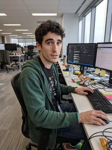 Ali, AI character: a quiet Toronto software office QA corner, two monitors on a desk, snack wrappers and sticky notes around…