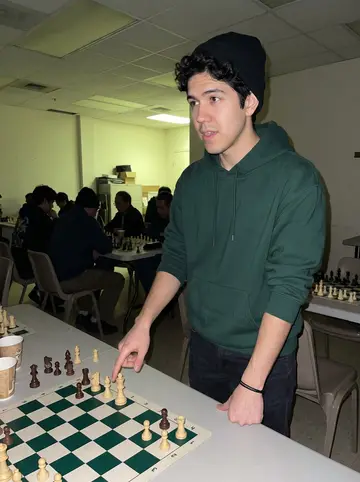 Amir, AI character: back room of a late-night community center chess meetup, folding tables, paper cups and several boards…