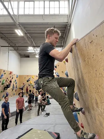 Jerry, AI character: an indoor rock climbing gym in SoMa, halfway over the top of a bouldering wall with colored holds and…