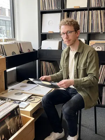 Kaleb, AI character: a small vinyl shop listening corner near the used records bins, blank record sleeves stacked on a low table