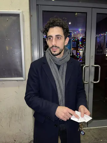Hassan, AI character: outside a cheap late showing at a Manchester cinema, holiday lights reflected in the glass doors