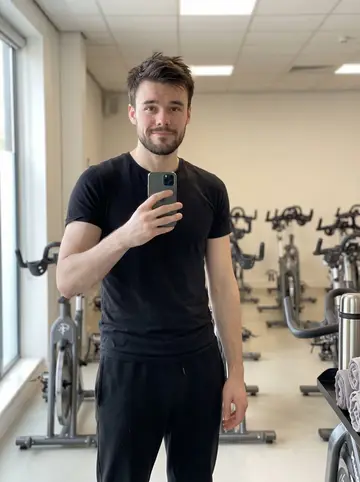 Orlando, AI character: the bright cycling room at the Brighton fitness club just after his dawn spin class, leaning beside a…