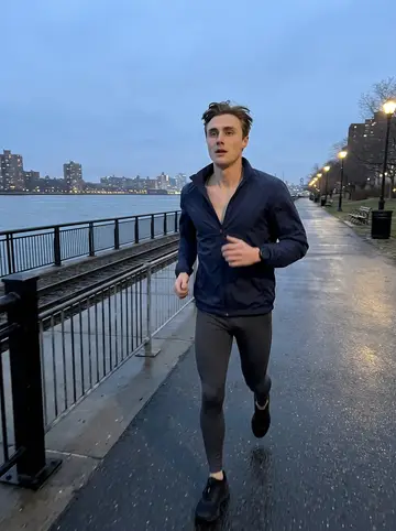 Tanner, AI character: the East River running path before sunrise, wet pavement, black railing, Queens lights across the water