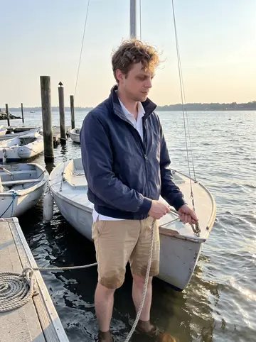 Forrest, AI character: a small sailing club dock on the Connecticut coast near Greenwich, dinghies tied up behind him