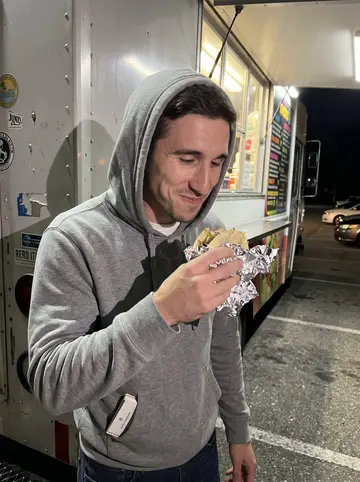 Alec, AI character: beside a taco truck in a Savannah parking lot at night, menu board glow and parked cars behind him