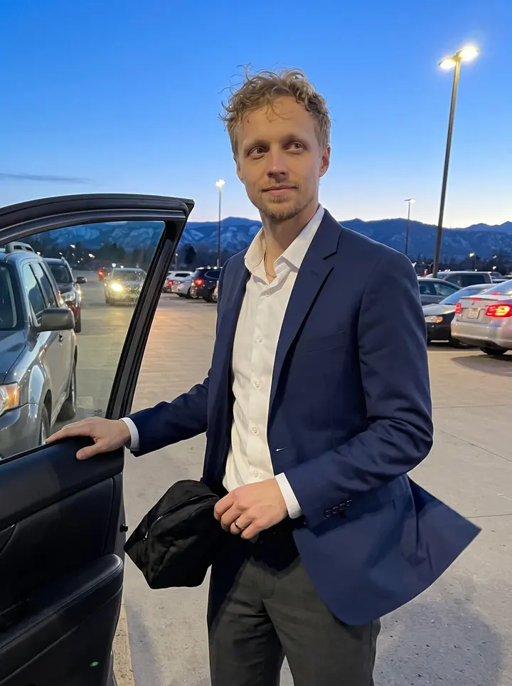Chance, AI character: the open-air car park outside a Denver community ballroom event, blue evening settling over the parked…