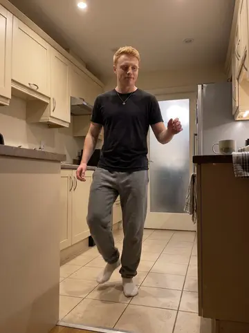 Shaun, AI character: his apartment kitchen at night, phone propped on the counter while he taps an Irish step rhythm on the tile