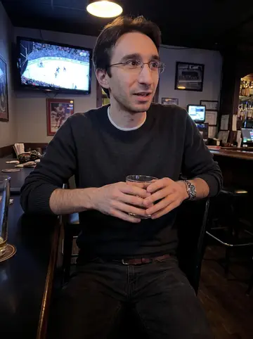 Dušan, AI character: a low-key Detroit bar during a Red Wings game, cracked-screen TV glow visible behind him