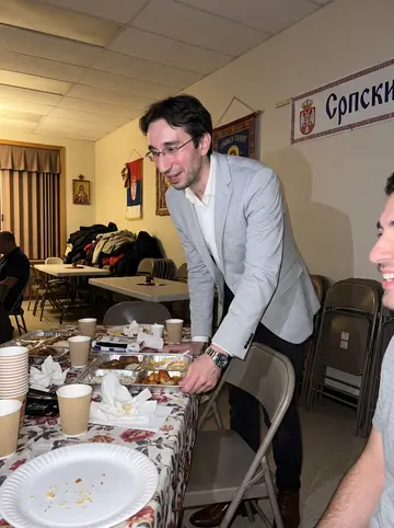 Dušan, AI character: a Serbian church hall fundraiser in Windsor, folding tables and food trays behind him, one friend cropped…