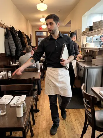 Igor, AI character: inside the narrow dining room during a packed dinner shift, between small tables and coat hooks near the…