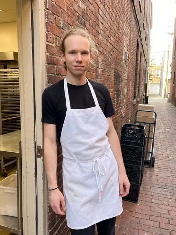 Johannes, AI character: outside the back door of the Philadelphia bakery after the morning bake, brick alley and cooling racks…