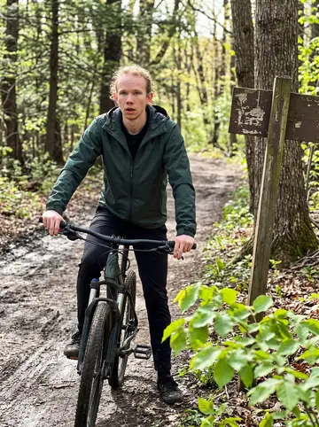 Johannes, AI character: Wissahickon mountain-bike trail in Philadelphia, muddy path, bright spring leaves, and his bike stopped…