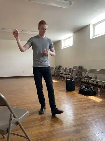 Lukáš, AI character: church basement practice hall with a polished floor, folding chairs against the wall and his black dance…