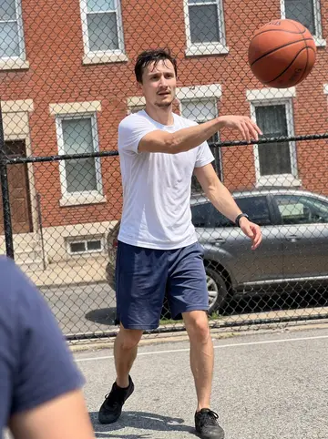 Paweł, AI character: outdoor pickup basketball court in Fishtown, chain-link fence and brick rowhouses behind him