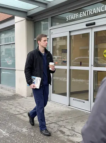 Aleksi, AI character: outside the staff entrance of a Seattle hospital between cases, cold clear morning pavement and glass…