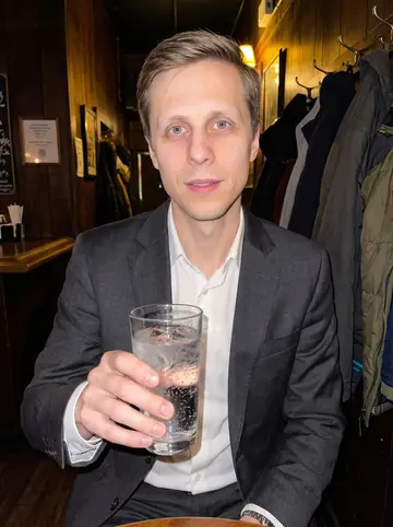 Aleksi, AI character: a low-key hospital fundraiser after-party at a narrow Seattle bar, wood wall and coats behind him