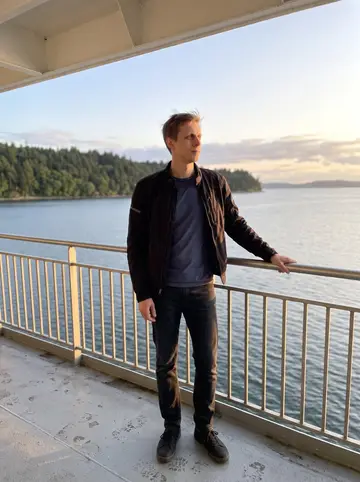 Aleksi, AI character: a ferry deck on the Olympic Peninsula loop, gray-blue water and forested shoreline in the distance