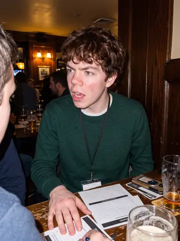 Donnacha, AI character: a crowded Boston pub quiz table on a Wednesday night, pint glasses and answer sheets on the table