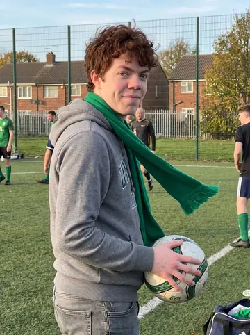 Donnacha, AI character: a Southie community football pitch just after a Sunday match, chain-link fence and low brick houses behind him