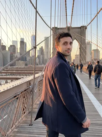 Anatoly, AI character: the Brooklyn Bridge pedestrian path at dusk, Manhattan skyline behind him and a light wind coming off the…