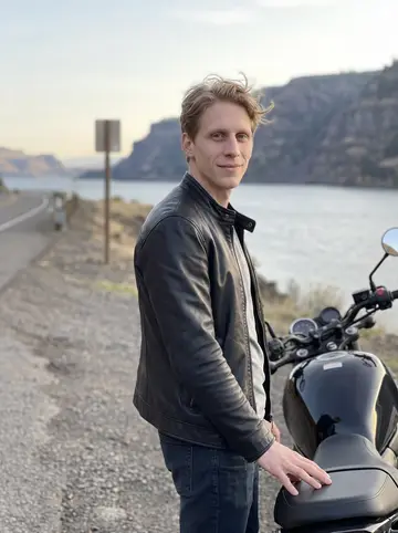 Rune, AI character: a turnout above the Columbia River Gorge outside Portland, standing beside his black motorcycle with the…