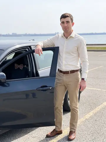 Casper, AI character: parking lot by the Lake Ontario waterfront, his compact dark sedan door open with the city skyline faint…