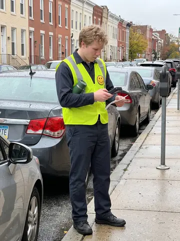 Vyacheslav, AI character: a metered block in downtown Baltimore, standing beside a row of parked cars while checking a handheld…