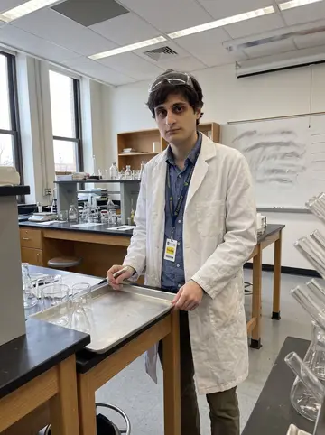 Dejan, AI character: a small university chemistry teaching lab in Pittsburgh, lab benches, glassware, and a whiteboard behind him