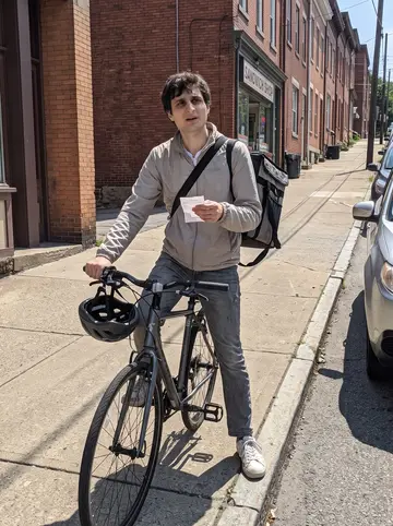 Dejan, AI character: outside a sandwich shop on a steep Pittsburgh street, bike locked beside the curb and row houses climbing…