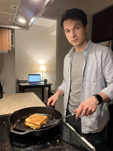 Veljko, AI character: his Toronto apartment kitchen after midnight, laptop open on the table and a cast-iron pan on the stove