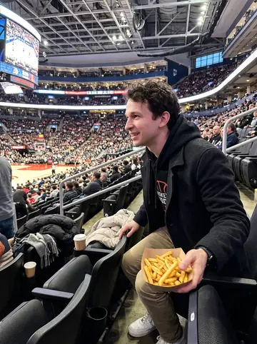 Veljko, AI character: upper-bowl cheap seats at a Raptors game in Toronto, court lights far below and winter coats piled on the…