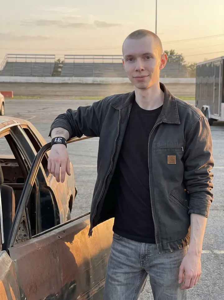Jukka, AI character: outside the Oshawa dirt oval at golden hour, standing beside his rusted stock car with the pit lane and…