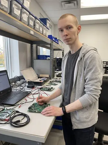 Jukka, AI character: electronics test bench at his firmware job in Oshawa, small circuit boards, cables, a laptop, and a shelf…