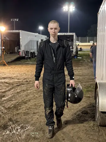 Jukka, AI character: Saturday night dirt oval pit area after a stock car heat, damp clay on the ground, trailers and track…