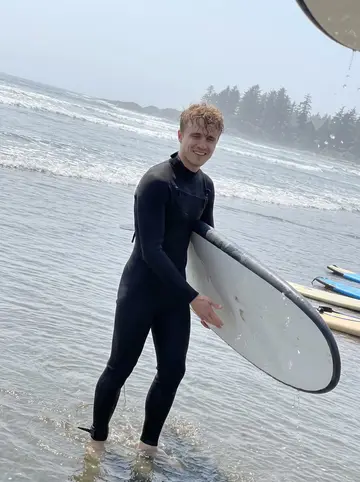 Ilari, AI character: a beginner surf lesson beach near Tofino, grey-blue Pacific water and rental boards on the sand