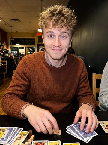 Ilari, AI character: a crowded board game night at a neighborhood cafe in Mount Pleasant, cards and wooden pieces on the table