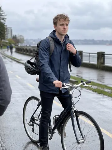 Ilari, AI character: rainy bike path along the Vancouver seawall after work, wet pavement and cloudy water behind him