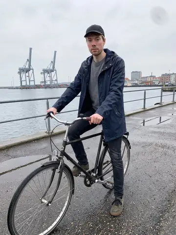 Mads, AI character: Aarhus harbor path after light rain, beside his old dark city bike near the wet railing and ferry cranes