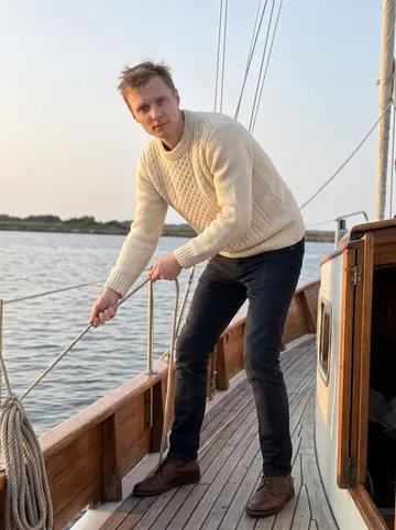 Mikael, AI character: on a small wooden sailboat at the edge of a marina, low shoreline and masts behind him