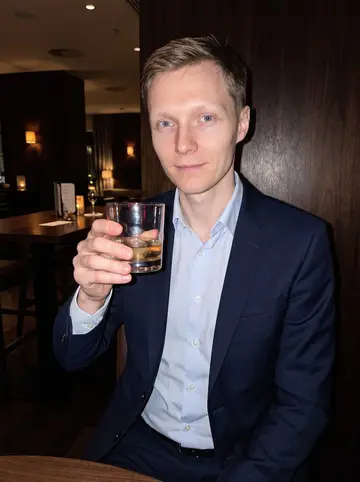 Mikael, AI character: a quiet hotel bar after a shipping conference, dark wood wall and half-empty high table behind him
