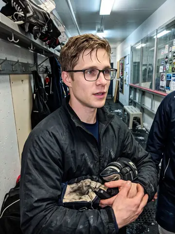 Heikki, AI character: local ice rink hallway after Thursday-night amateur hockey with the Cavities, rink boards and skate racks…
