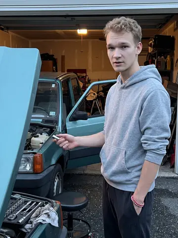 Teemu, AI character: shared garage driveway beside his brother's old Fiat Panda, hood open, socket set on a rolling stool