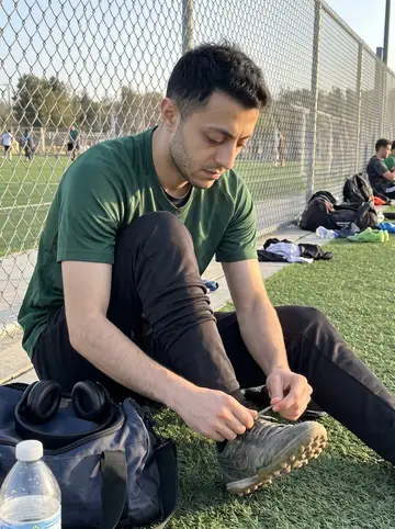 Mehdi, AI character: Thursday evening pickup soccer at an Irvine rec field, chain-link fence and green turf behind him