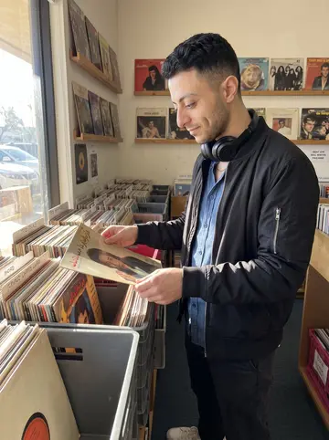 Mehdi, AI character: a used vinyl shop in Orange County, narrow aisles of record crates and a sunlit front window behind him