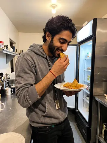 Dhruv, AI character: the motel back kitchen late at night, with a paper plate of samosas on a stainless counter and a half-open…