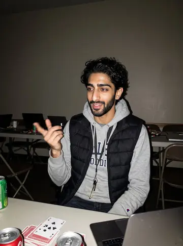 Dhruv, AI character: a campus lounge during a winter coding-club game night, folding table with cards, soda cans and laptops…
