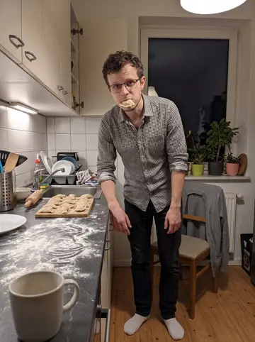 Łukasz, AI character: his Berlin apartment kitchen, crowded with hardy houseplants on the windowsill and a small board of uneven…