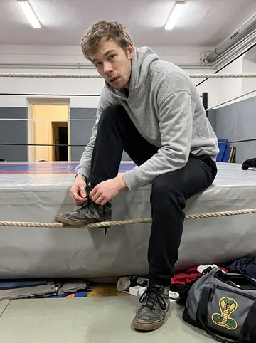 Štěpán, AI character: a small wrestling gym in Favoriten after Thursday night practice, ring ropes and padded floor behind him
