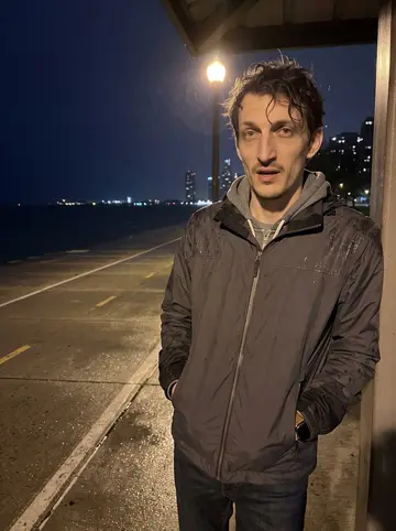 Dariusz, AI character: the Chicago lakefront path after rain, dark water and wet pavement behind him, city lights blurred but…